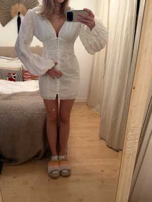 Studentklänning från adoore - Söt vit miniklänning med diskreta prickar, djup V-ringning och långa puffärmar. Klänningen har knappar framtill och markerad midja som ger en feminin siluett. Tillverkad i ett lätt, strukturerat tyg som känns luftigt och fräscht. Detta mönster säljs ej längre och klänningen har inga defekter. Storlek 40 men passar en 38 💕