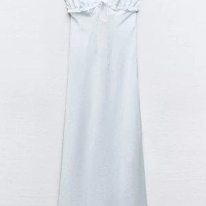 Vit långklänning i satin från Zara - Supersnygg vit långklänning från Zara i glansig satin med smala axelband, v-ringning och söt snörning framtill. Klänningen har volangdetaljer vid bysten och en elegant, flowy passform. Perfekt för dig som vill sticka ut med en stilren look.