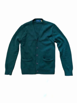 Polo Ralph Lauren Cardigan - Tja! Säljer nu en stilren mörkgrön cardigan från Polo Ralph Lauren i otroligt fint skick,  fem knappar framtill och i frägen mörkgrön. Nypris ca 3000 kr, säljer för knappt halva! Två fickor framtill och ribbade muddar vid ärmslut och nederkant. Perfekt för lager på lager och enkel att matcha. Storlek M. Pris går alltid att diskuteras✅. 