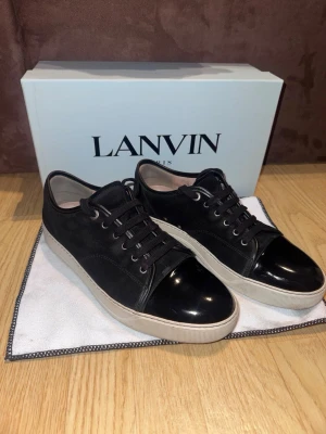Svarta Lanvin  - Snygga svarta sneakers från Lanvin med klassisk låg siluett. Skorna har en unik blank lackad tå, svarta skosnören och vit sula. Ovandelen är i mocka och textil vilket ger en lyxig känsla. Perfekta för dig som vill ha något stilrent men ändå sticker ut. Uk 9