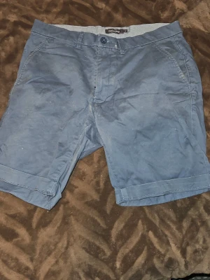 Blåa shorts från Dressmann - Snygga blåa shorts från Dressmann med klassisk passform och knappgylf. Shortsen har bälteshällor, sidofickor och är tillverkade i ett mjukt bomullsmaterial. Perfekta för varma dagar och enkel att matcha med olika stilar.