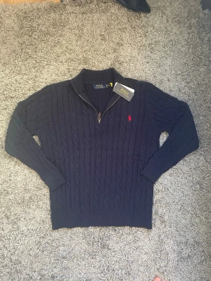 Mörkblå kabelstickad tröja Polo Ralph Lauren - Säljer en mörkblå kabelstickad tröja från Polo Ralph Lauren med halv dragkedja och ribbade muddar. Tröjan har det klassiska röda Polo-logot broderat på bröstet och är tillverkad i mjukt material. Perfekt för dig som gillar stilren och tidlös design.