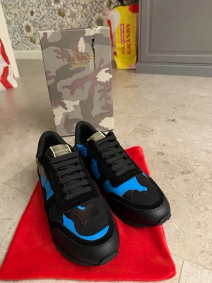 Valentino Garavani svarta/blå sneakers - Snygga sneakers från Valentino Garavani i svart och blått med coolt camomönster. Skorna har svarta snören, gummisula och detaljer i läder och textil. Perfekta för dig som vill sticka ut med en exklusiv look och streetwear-vibe.