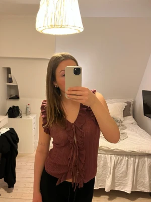 Vinröd volangtopp med knyt - Snygg vinröd topp med volangdetaljer längs axlar och framkant. Toppen har v-ringning och knyt framtill, är ärmlös och har en lätt ribbad struktur. Perfekt för dig som gillar detaljer och en trendig look.