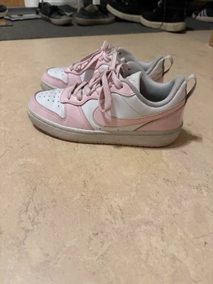 Nike rosa och vita låga sneakers - Säljer ett par snygga Nike sneakers i lågt modell med rosa och vita paneler. Skorna har klassisk Nike Swoosh på sidan, vita snören och grå detaljer vid häl och insida. Perfekta för dig som gillar en clean och sportig look.