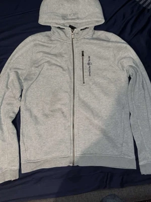 Ljusgrå hoodie från Sail Racing - Ljusgrå hoodie från Sail Racing med dragkedja hela vägen, bröstficka med dragkedja och broderad logga på bröstet. Huva med dragsko och diskret logga även på huvan. Tillverkad i mjukt bomullsmaterial, perfekt för chill dagar.