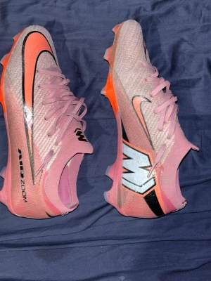 Elite Nike rosa Mercurial fotbollsskor - Säljer ett par Nike Elite Mercurial fotbollsskor i rosa med svarta och orange detaljer. Skorna har en stor svart Swoosh-logga, AIR ZOOM-tryck på sidan och en cool M-detalj på hälen. Ovandelen är i syntetmaterial och sulan har fasta dobbar för spel på gräs. Storlek 41/42