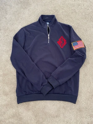 Jeffrey Epstein Quarter Zip Bomull - Jeffrey Epsteins Island tröja med halv dragkedja och ståkrage. På bröstet finns ett rött broderat monogram och på ena ärmen sitter en stor amerikansk flagga med gul kant. Tröjan har ribbade muddar vid ärmslut och nederkant. Perfekt för dig som gillar stilrena detaljer och vill sticka ut.