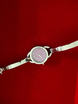 Silverfärgad klocka med lila urtavla - Stilren klocka med smalt silverfärgat armband och rund boett. Urtavlan är lila med diskreta ränder och små silverprickar som timmarkeringar. Klassisk quartz-urverk och enkel design som passar till många stilar.