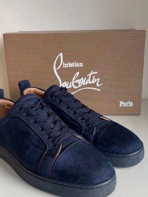 Mörkblå sneakers från Christian Louboutin - Snygga mörkblå sneakers i mocka från Christian Louboutin. Skorna har svarta snören, diskreta sömmar och en klassisk låg siluett. Yttersulan är svart och insidan är fodrad i beige läder för extra komfort. Perfekta för dig som gillar exklusiv och stilren design.