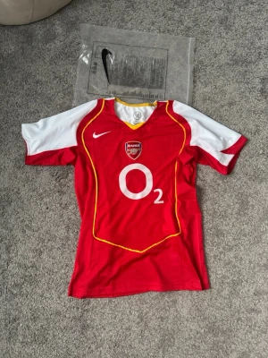 Arsenal Nike matchtröja röd/vit - Säljer en klassisk Arsenal matchtröja från Nike i rött och vitt med gula detaljer. Tröjan har klubbmärke och O2-logga på bröstet samt korta ärmar. Perfekt för dig som älskar fotboll och vill ha en ikonisk supportertröja i sportigt material.