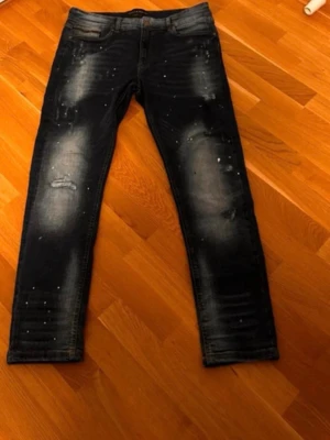 Mörkblå slitna jeans med dragkedjedetalj - Säljer ett par mörkblå jeans med slitningar och coola färgstänk. Jeansen har skinny fit och en unik dragkedjedetalj vid fickan. Materialet är klassisk denim med stretch. Perfekta för dig som gillar en trendig och avslappnad stil.