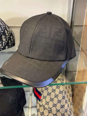 Svart keps från Fendi med ton-i-ton-mönster - Snygg svart keps från Fendi med diskret ton-i-ton FF-logomönster över hela kepsen. Böjd skärm med läderdetalj längs kanten ger en exklusiv känsla. Perfekt accessoar för dig som gillar stilrena designer och lyxiga detaljer. Kommer med box och dustbag men då kostar det 400 extra eftersom allt är äkta
