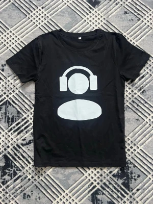 Svart t-shirt med hörlursmotiv - Cool svart t-shirt med ett stort vitt tryck av en figur med hörlurar på bröstet. Klassisk rund halsringning och korta ärmar. Perfekt för dig som gillar musik och vill ha en enkel men uttrycksfull stil. Tillverkad i mjuk bomull för skön känsla.