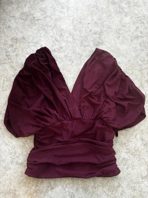 Vinröd croppad topp med puffärm - Trendig vinröd croppad topp med djup v-ringning och stora puffärmar. Toppen har en smickrande passform och markerad midja, perfekt för att lyfta vilken outfit som helst. Vet ej vart den är ifrån då de inte finns nån lapp..
