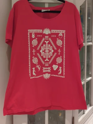 Rosa t-shirt med tribaltryck  - Säljer en rosa t-shirt från Bluewear Woman med vitt tribal-inspirerat tryck framtill. T-shirten har rund halsringning och korta ärmar. Materialet känns mjukt och bekvämt, perfekt för en avslappnad stil.