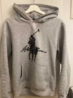 Polo hoodie - Snygg grå hoodie från Polo Ralph Lauren med stor svart polospelare och signatur på bröstet. Klassisk känguruficka framtill och mjukt material i bomullsblandning. Perfekt för en chill och stilren look.