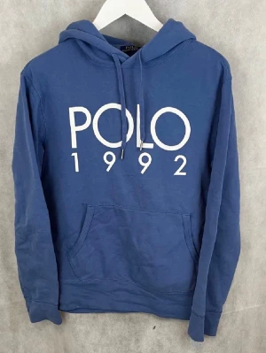 Polo hoodie - Blå hoodie från Polo Ralph Lauren med stor vit POLO 1992-print på bröstet. Klassisk huva med dragsko och känguruficka framtill. Tillverkad i mjukt bomullsmaterial, perfekt för en avslappnad och stilren look.