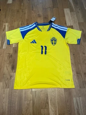 Sweden 2026 home kit X elanga #11 - Gul och blå Sverige matchtröja från Adidas med Elanga och nummer 11 på ryggen. Tröjan har korta ärmar, broderad landslagssköld och Adidas-logga på bröstet. Tillverkad i lätt och ventilerande material, perfekt för fotboll eller träning.