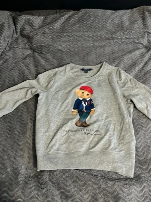 Grå sweatshirt Polo Ralph Lauren Bear - Grå sweatshirt från Polo Ralph Lauren med ikoniska Polo Bear-trycket framtill. Det är barn storlek, enligt guide så passar den 144-154. Den är i fint skick. Pris och frågor är det bara att höra av sig!