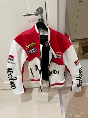Röd och vit Marlboro racing skinnjacka - Säljer en riktigt snygg racinginspirerad skinnjacka från Marlboro i rött och vitt med svarta detaljer. Äkta läder, några svarta streck som var där när jag köpte den (ny). Mycket bra skick och den är fodrad så hyfsat varm. Herr L, sitter oversized. Kan sänka pris vid snabb affär 