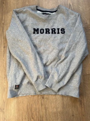 Grå sweatshirt från Morris - Säljer en grå sweatshirt från Morris med svart broderad logga på bröstet. Tröjan har rund halsringning, långa ärmar och ribbade muddar vid ärmslut och nederkant. Perfekt för en avslappnad och stilren look.