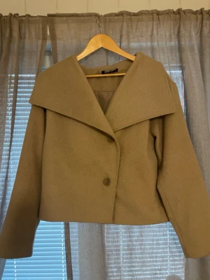 Beige kappa från Gina Tricot - Stilren beige kappa från Gina Tricot med bred krage och två knappar framtill. Kortare modell med raka ärmar och mjukt material som ger en snygg siluett. Perfekt för dig som gillar minimalistisk och trendig look.