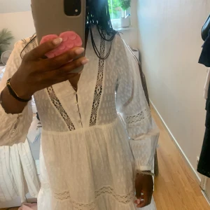 Vit kort klänning med spetsdetaljer - Superfin vit kortklänning med v-ringning, långa ärmar och spetsdetaljer längs bröstet, ärmarna och kjolen. Klänningen har knappar framtill och ett lätt, luftigt tyg med diskret prickmönster. Perfekt för sommarens alla tillfällen!💞💞💞