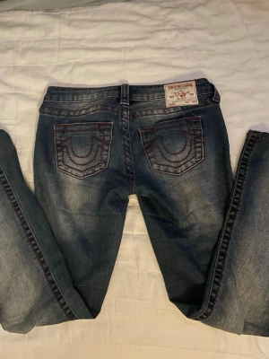 True Religion jeans W25🤍 - Säljer ett par blå jeans från True Religion med ikoniska röda kontrastsömmar och broderade bakfickor. Jeansen har en klassisk femficksmodell och är tillverkade i denim med lätt tvättad look. Dem är lite uppsydda av tidigare ägare✨✨