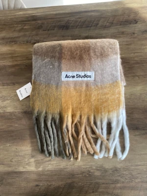 Rutig halsduk från Acne Studios - Säljer en fluffig och mjuk halsduk från Acne Studios i rutigt mönster med färgerna beige, brunt, gult, vitt och grått. Halsduken har långa fransar i ändarna och en stor Acne Studios-logga. Perfekt för att addera färg till din outfit.