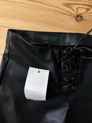 Svarta utsvängda byxor från Zara - Snygga svarta byxor i fuskläder från Zara med utsvängda ben och snörning framtill i midjan. Byxorna har en modern look och passar perfekt till dig som vill sticka ut med en trendig stil.