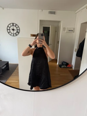 Svart halterneck klänning omlott - Säljer en svart kort halterneck klänning med omlott och öppen rygg. Klänningen har en lös passform och knyts i nacken. Perfekt för sommaren och enkel att styla med accessoarer.