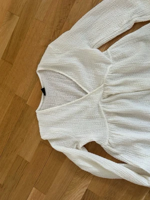 Vit omlottklänning från Gina Tricot - Säljer en vit omlottklänning från Gina Tricot i strukturerad bomull. Klänningen har lång ärm, v-ringning och ett lätt rynkat liv som ger en snygg siluett. Perfekt för dig som gillar en enkel men stilren look.