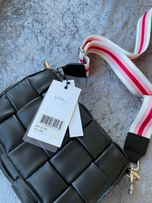 Svart flätad axelväska med bred rem - Trendig svart axelväska i flätad konstläder med bred, justerbar axelrem i vitt och rosa. Väskan har magnetlås och silverfärgade detaljer. Perfekt för dig som vill ha en cool och modern accessoar till din outfit.Det rosa bandet är bara ett extra band ni får med, det riktiga finns i väskan . Innerdelen kan man ta loss om man önskar 