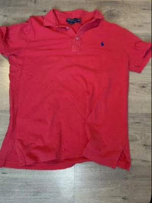Röd pikétröja från Polo Ralph Lauren - Klassisk röd pikétröja från Polo Ralph Lauren med blå broderad logga på bröstet. Tröjan har korta ärmar, krage och knappar vid halsen. Perfekt till jeans eller shorts för en avslappnad men snygg look.