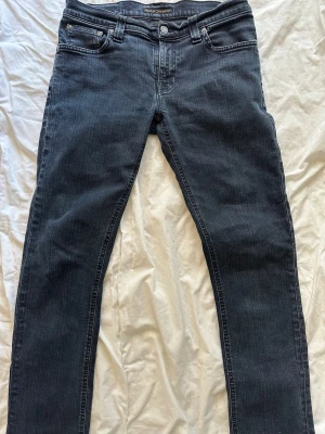Mörkblå jeans från Nudie Jeans - Säljer ett par mörkblå jeans från Nudie Jeans med klassisk femficksmodell och raka ben. Jeansen har silverfärgade nitar och knapp, samt diskreta slitningar för en snygg look. Perfekta till en avslappnad stil. Storlek:29:34