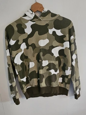 Camo hoodie med huva - Snygg hoodie med camouflagemönster i grönt, beige och vitt. Tröjan har en stor ficka framtill och en klassisk huva. Perfekt för dig som gillar streetwear och vill sticka ut lite extra. Mjuk och skön bomullsblandning.