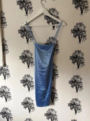 Blå satinklänning med smala axelband - Supersnygg blå satinklänning med smala axelband och figurnära passform. Klänningen har en kort längd och en enkel, stilren design som ger en cool och trendig vibe. Perfekt för dig som vill sticka ut med en glansig look.