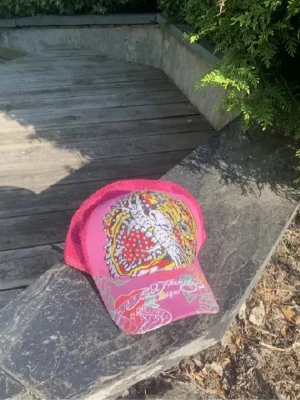 Rosa Ed Hardy keps med tigertryck - Snygg rosa keps från Ed Hardy med ikoniskt tigertryck framtill, dekorerad med glittrande stenar. Kepsen har mesh-bakstycke för extra ventilation och justerbar passform. Perfekt för dig som gillar streetstyle och färgstarka accessoarer.