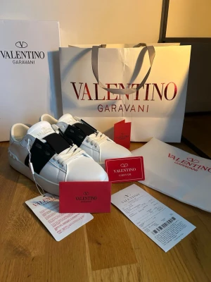 Valentino Opens - Snygga vita sneakers från Valentino Garavani med svart band över vristen och ikoniska nitar på hälen. Skorna har en klassisk låg siluett, vita snören och är tillverkade i slätt läder. Perfekta för dig som vill ha en exklusiv och trendig look. Obs kan gå ner i pris vid snabb affär! 