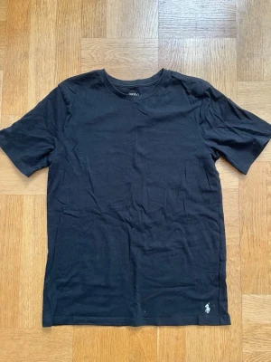 Svart t-shirt från Polo Ralph Lauren - Klassisk svart t-shirt från Polo Ralph Lauren med rund halsringning och korta ärmar. Liten broderad logga i vitt nertill på framsidan. Tillverkad i mjuk bomull för en skön känsla och enkel stil.