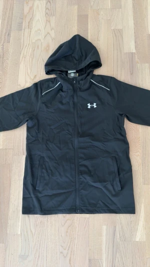 Svart vindjacka från Under Armour - Svart vindjacka från Under Armour med huva, hel dragkedja och två sidofickor. Jackan har vitt logotryck på bröstet och reflekterande detaljer vid axlarna. Tillverkad i lätt och funktionellt material, perfekt för löpning och träning utomhus.