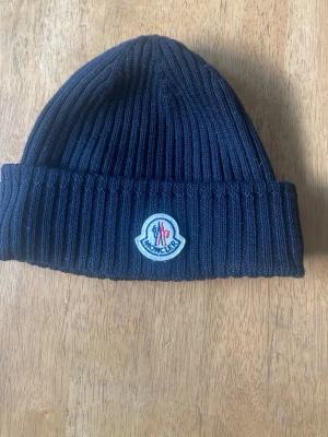 Svart ribbad mössa från Moncler - Säljer en svart ribbstickad mössa från Moncler med klassisk logotyp framtill. Mössan är i mjukt material och har en uppvikt kant för extra värme och stil.