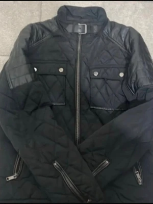 Svart pufferjacka från Zara Man - Snygg svart pufferjacka från Zara Man med quiltat mönster, dragkedja framtill och två bröstfickor med knappar. Jackan har detaljer i läderimitation på axlar och ärmar samt spänne vid kragen. Perfekt för kalla vinterdagar.