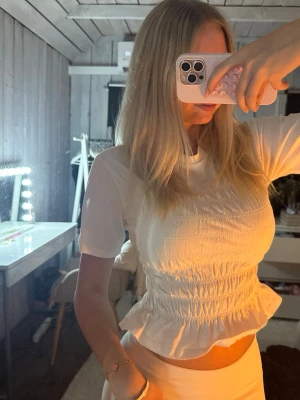 Vit smockad topp med volangkant - Supersöt vit topp med smockad midja och volangkant nedtill. Kortärmad modell med rund halsringning och figurnära passform. Perfekt till jeans eller kjol för en trendig look.