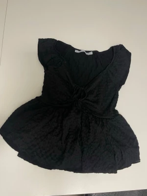 Svart blus med snörning i midjan - Supersöt svart blus med lätt struktur i tyget och snörning i midjan som ger en feminin siluett. Blusen har korta ärmar och en lätt v-ringad halsringning. Perfekt att styla med jeans eller kjol för en trendig look.