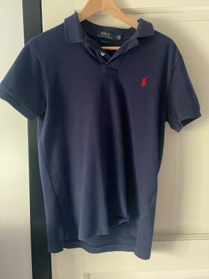Mörkblå piké från Polo Ralph Lauren - Klassisk mörkblå pikétröja från Polo Ralph Lauren med korta ärmar och röd broderad logga på bröstet. Tröjan har krage och knappar vid halsen, perfekt för en clean och stilren look. Tillverkad i mjuk bomull för skön komfort.