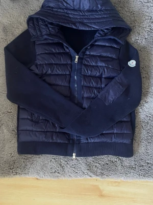 Mörkblå cardiganjacka från Moncler - Snygg mörkblå cardiganjacka från Moncler med quiltad front och huva. Ärmarna är stickade och har Moncler-logga på vänster arm. Jackan har dragkedja framtill och ribbade muddar. Perfekt för lager-på-lager och streetstyle.