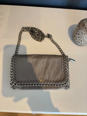 Grå axelväska med kedjedetaljer - Snygg grå axelväska med grov silverfärgad kedja som axelrem och dekor runt kanterna. Väskan har en mjuk, skrynklig läderlook och ett stilrent, rektangulärt format. Perfekt för dig som gillar en edgy och trendig stil.