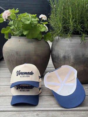 Voronado Riveira Cap - Snygg keps i beige med mörkblå skärm och broderad text 'Voronado Atelier' framtill. Kepsen har justerbar baksida och är tillverkad i ett klassiskt bomullsmaterial. Perfekt accessoar för en avslappnad och trendig stil.